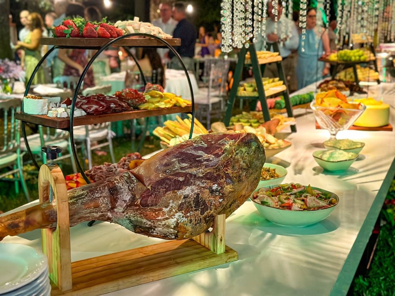 Mesa de buffet ao ar livre com pernil, frios, frutas e saladas em evento noturno