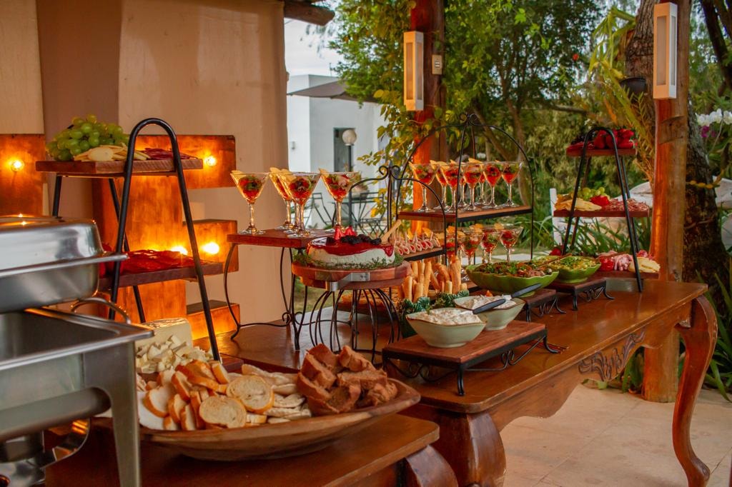 Mesa de buffet ao ar livre com taças, pães, saladas e iluminação quente ao entardecer