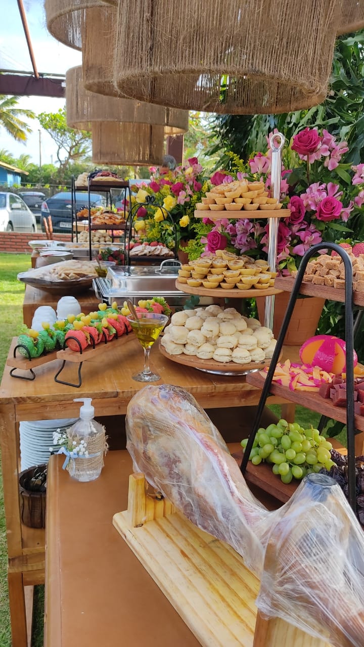 Mesa de buffet ao ar livre com mini-burgers coloridos, quiches, pães e flores rosas